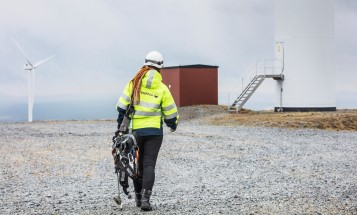 Image of a Vattenfall employee wearing safety clothes and helmet, walking across dry ground towards a wind turbine.